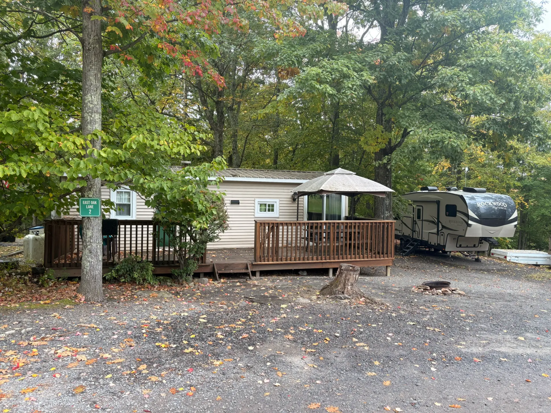 East Oak Lane 2 — East Oak Lane 2 with its long wooden deck, gazebo shade canopy, Rockwood RV parked beside it and a fire ring on a gravel site in the fall woods