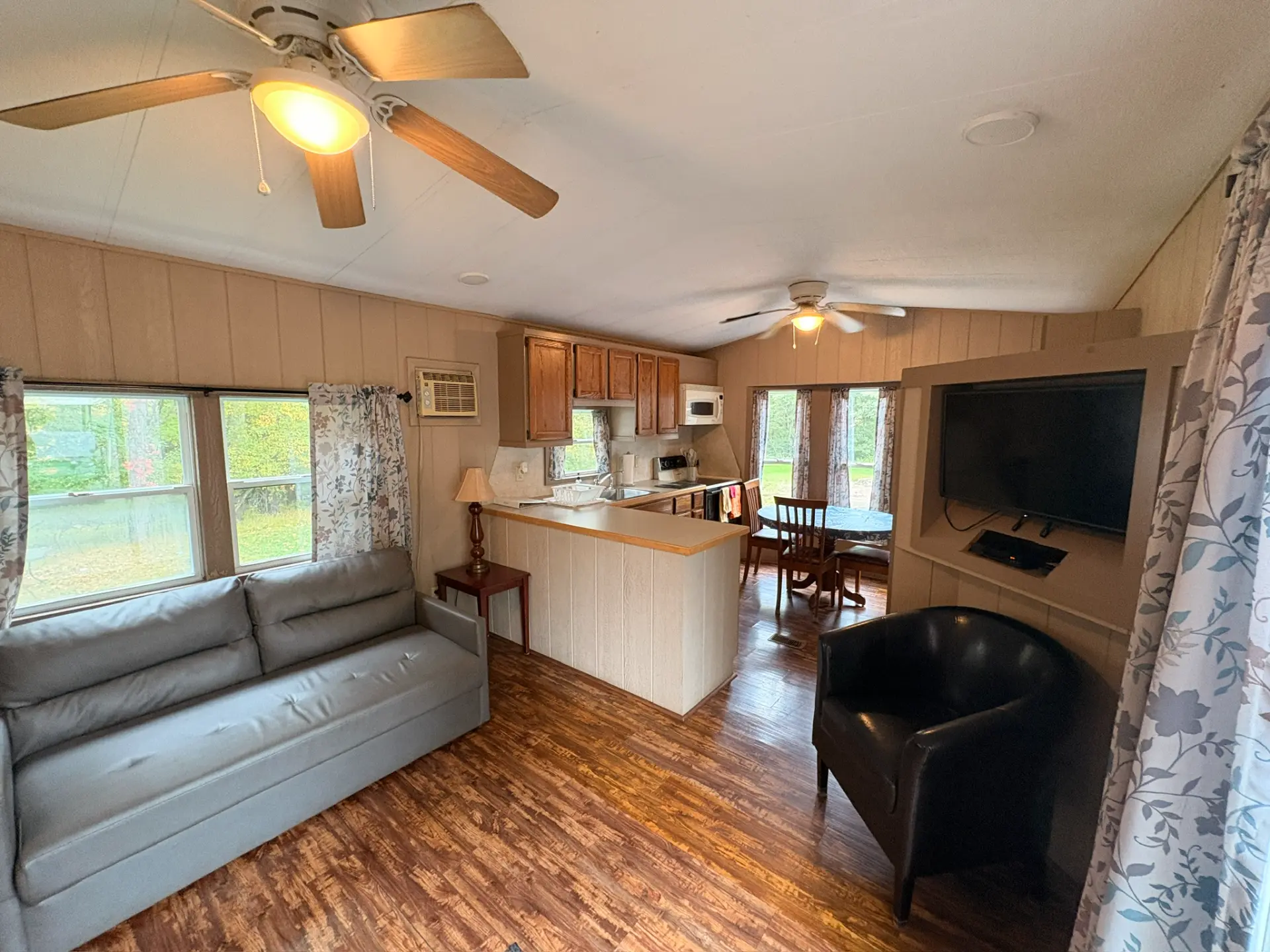 East Oak Lane 6 — Open living area and kitchen with a grey sofa under two windows, a black armchair by the wall-mounted TV, breakfast bar and oak cabinets