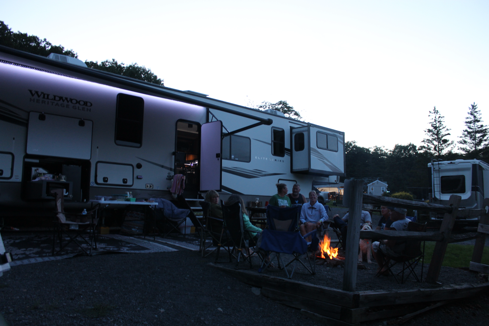 Fifth-wheel RV at a gravel site with family and friends gathered around a campfire at dusk