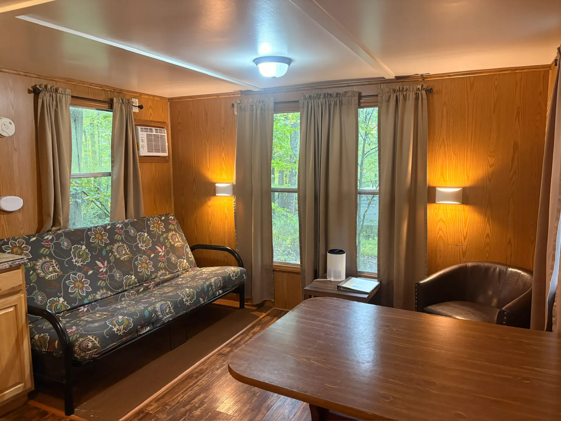 West Oak Lane 10 — Living area with a floral futon, dining table, leather chair, wall lamps and windows looking into the woods